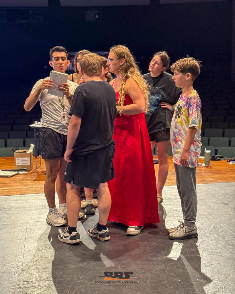 A group of students on a stage listening to a teacher.