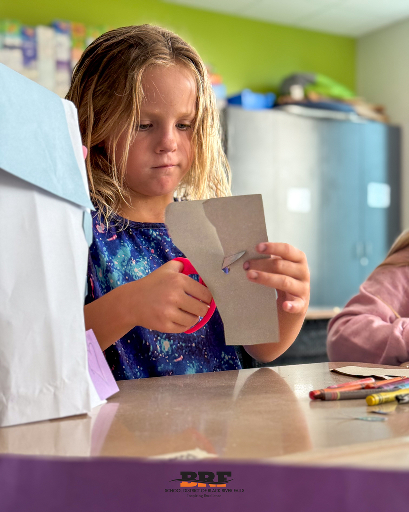 A young female student with light brown hair wearing a patterned blue shirt carefully uses pink safety scissors to cut a piece of gray cardboard at her desk. A paper bag project and crayons rest on the table beside her. School District of Black River Falls logo is at the bottom.