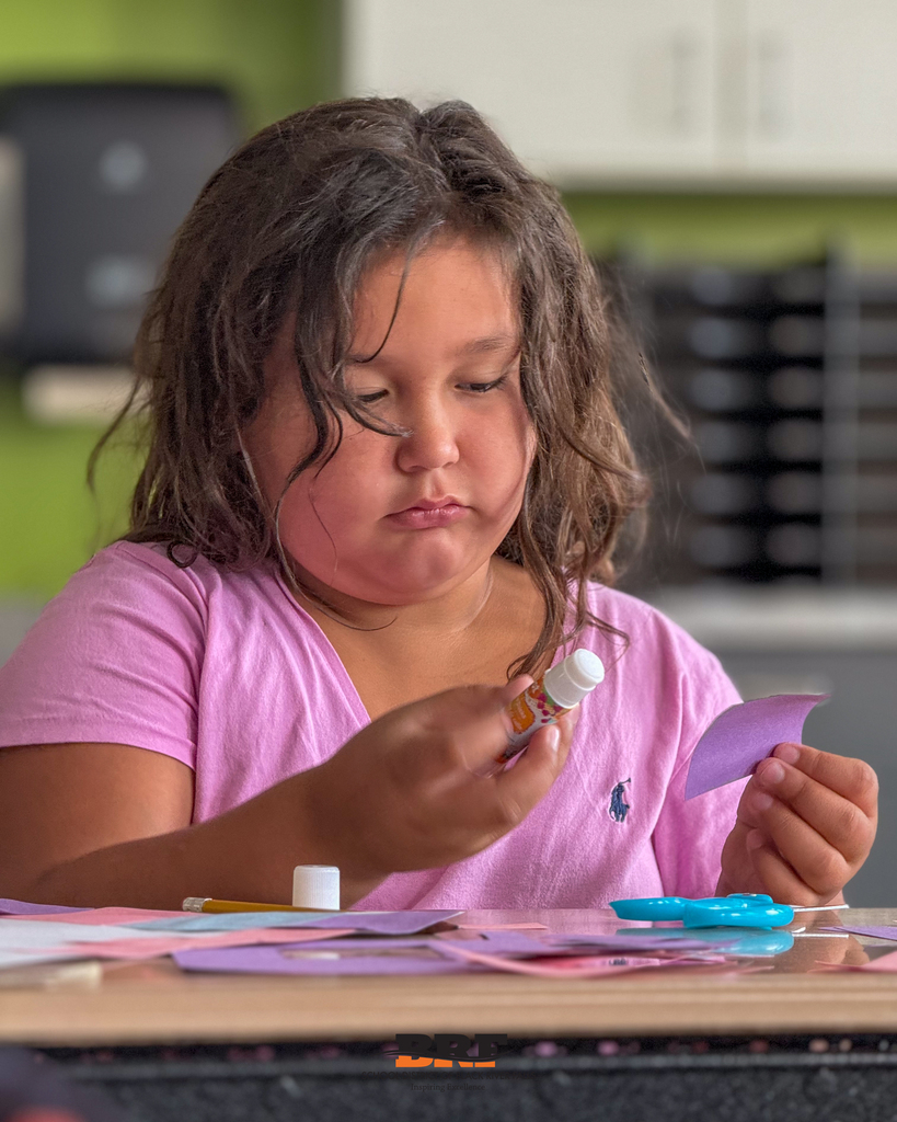 A young female student wearing a pink shirt sits focused at a classroom desk, carefully holding a glue stick in one hand and a piece of purple construction paper in the other during a crafting project. School supplies including scissors and paper scatter across the table. School District of Black River Falls logo is at the bottom.