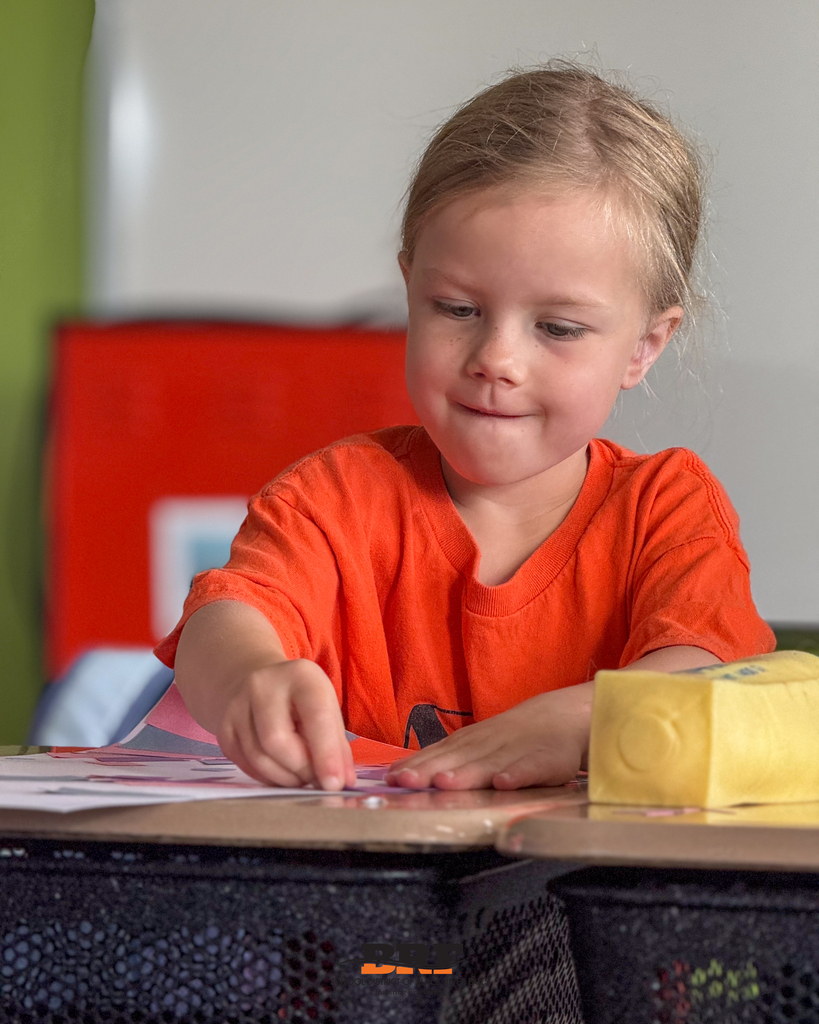 A young student wearing a bright orange shirt leans over a classroom desk with a focused smile while pressing colored paper onto a craft project. A pencil pouch sits on the desk nearby. School District of Black River Falls logo is at the bottom.
