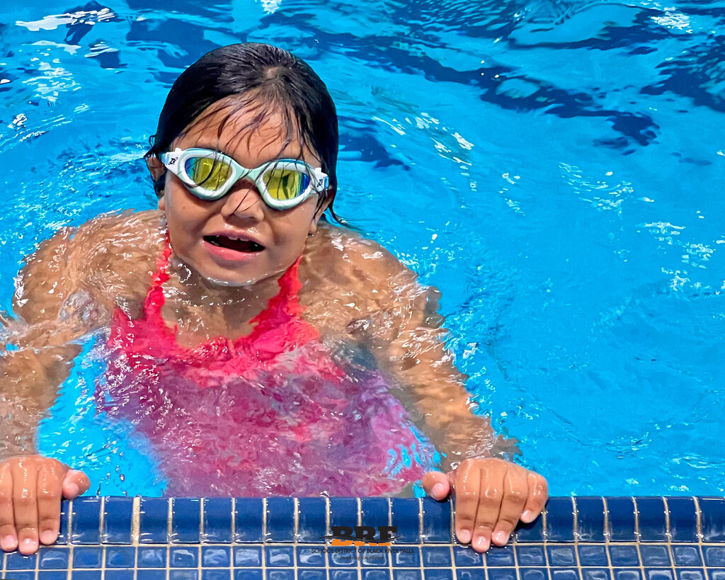 Girl in swimming pool grips side of pool with hands and smiles.