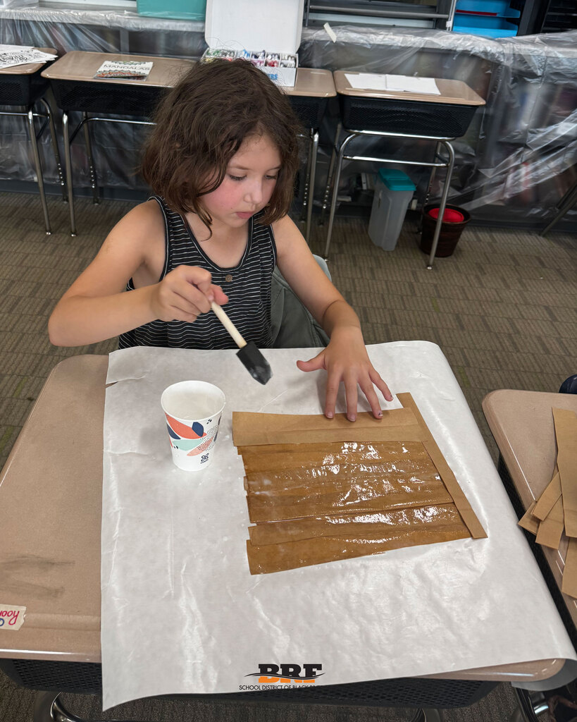 A young female student sits at her classroom desk using a black foam brush and liquid glue to seal horizontal strips of brown paper bag craft material on top of white paper. School District of Black River Falls logo is at the bottom.
