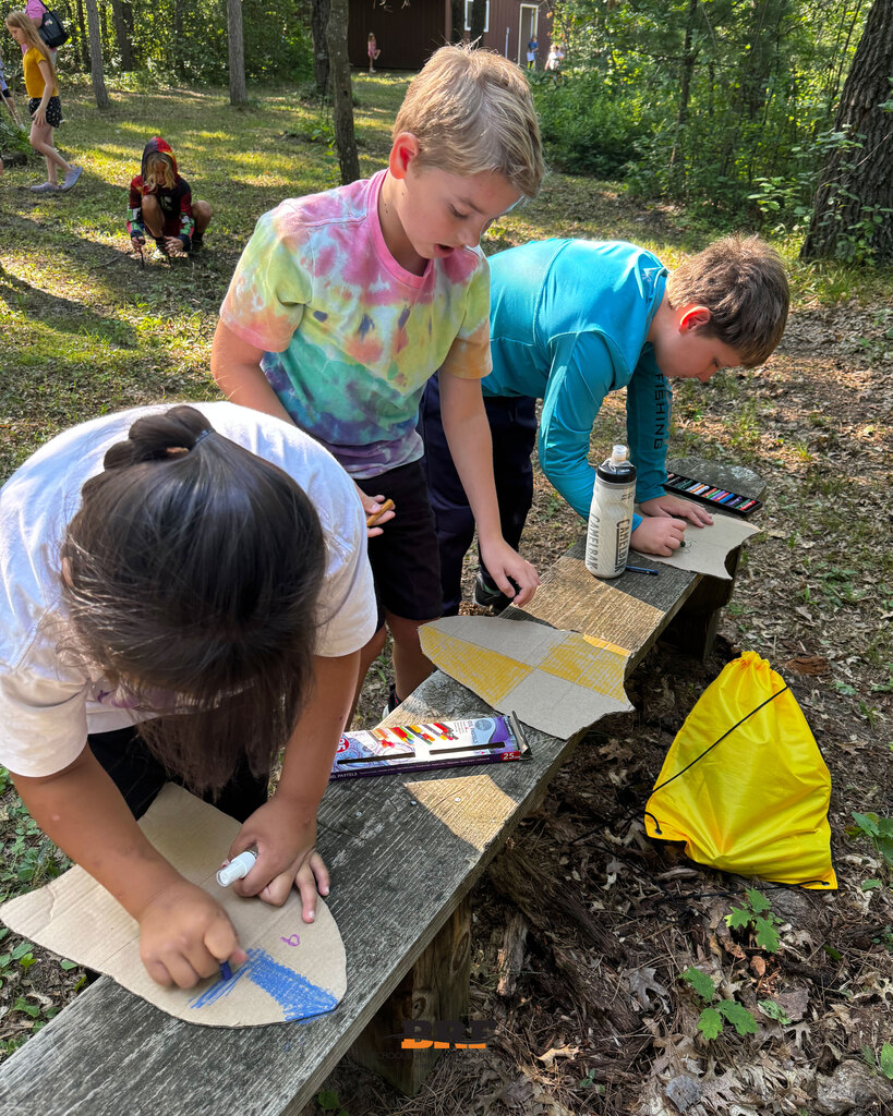 Three elementary students sit on a outdoor wooden bench in a wooded area during summer school. They are focused on drawing shields on cut-out cardboard using colorful pastels, with a set of oil pastels and a yellow cinch bag nearby. School District of Black River Falls logo is at the bottom.