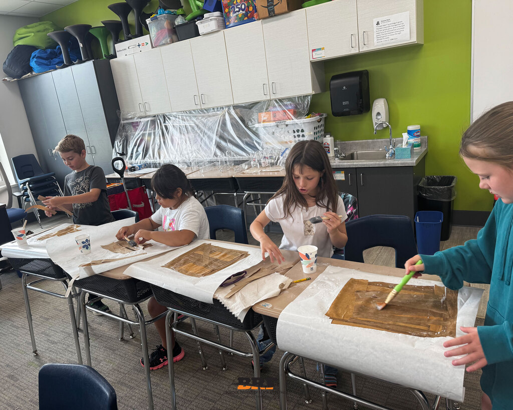 Four elementary students sit at classroom desks lined up side-by-side, using foam brushes and water-based glue to coat layered brown paper strips for a papyrus project. White protective paper covers the desks. School District of Black River Falls logo is at the bottom.