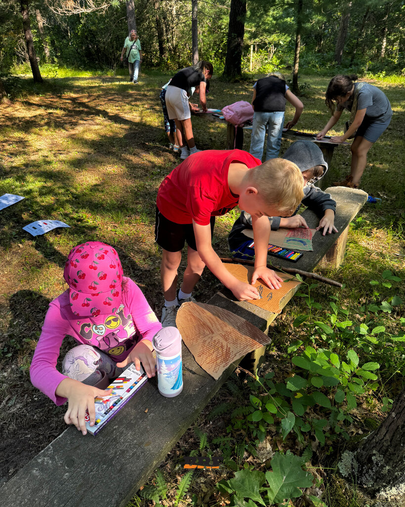 Three elementary students sit on an outdoor wooden bench in a wooded area during summer school. They are focused on drawing shields on cut-out cardboard using colorful pastels, with a set of oil pastels nearby. School District of Black River Falls logo is at the bottom.