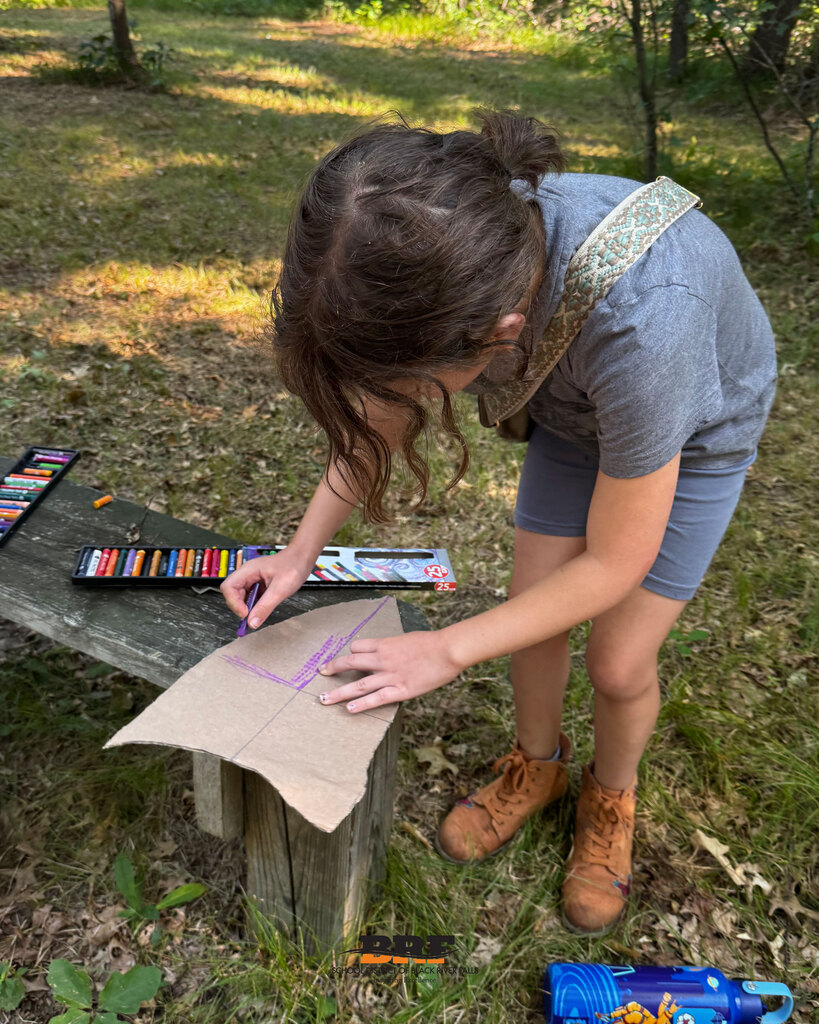 A young student with her hair tied back in a bun leans over an outdoor bench in a sunny forest setting, using a purple pastel to color a cardboard shield shape. A box of oil pastels rests on the wooden bench next to her. School District of Black River Falls logo is at the bottom.
