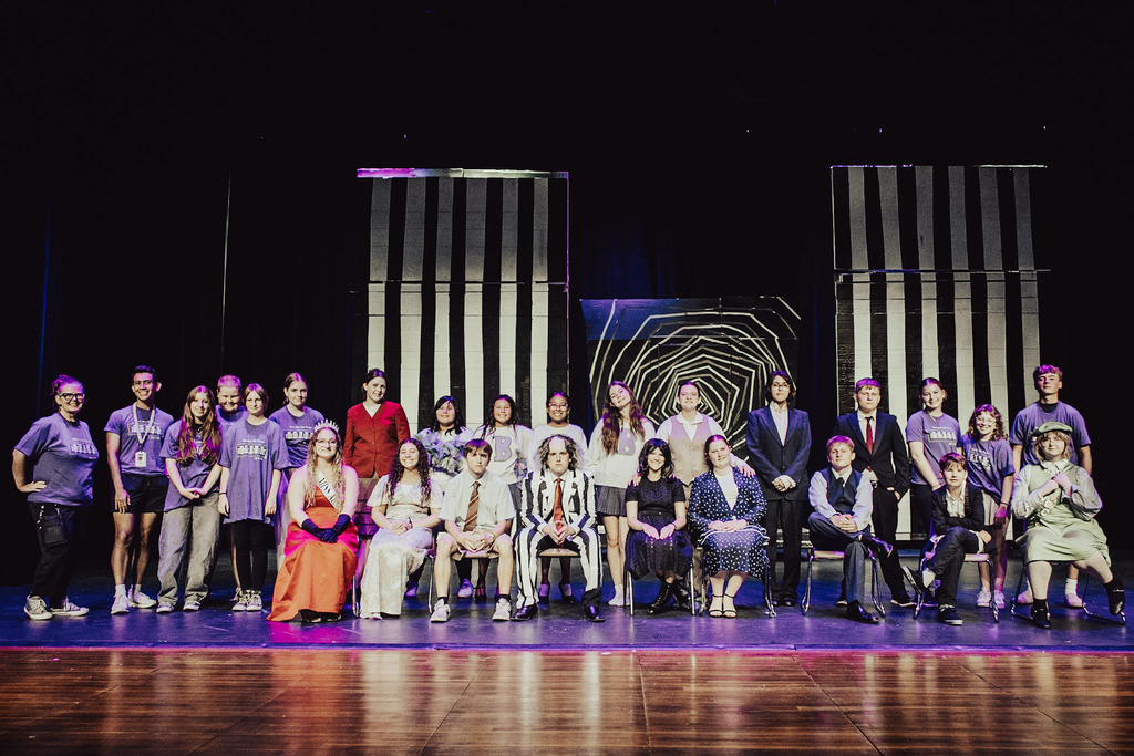 A group photo of the cast and crew of the middle school theater production of "Beetlejuice Jr." gathered on stage in the Lunda Theater. Students pose in full costume and show t-shirts in front of black-and-white striped set pieces. Sitting front and center is a student dressed as Beetlejuice in a signature black-and-white striped suit, flanked by cast members in theatrical costumes including a red gown, suits, and vintage dresses, while crew members in purple shirts stand along the sides.