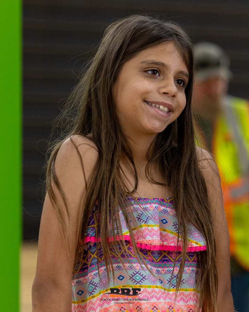 A young girl with long brown hair wearing a colorful patterned sundress, smiling warmly.