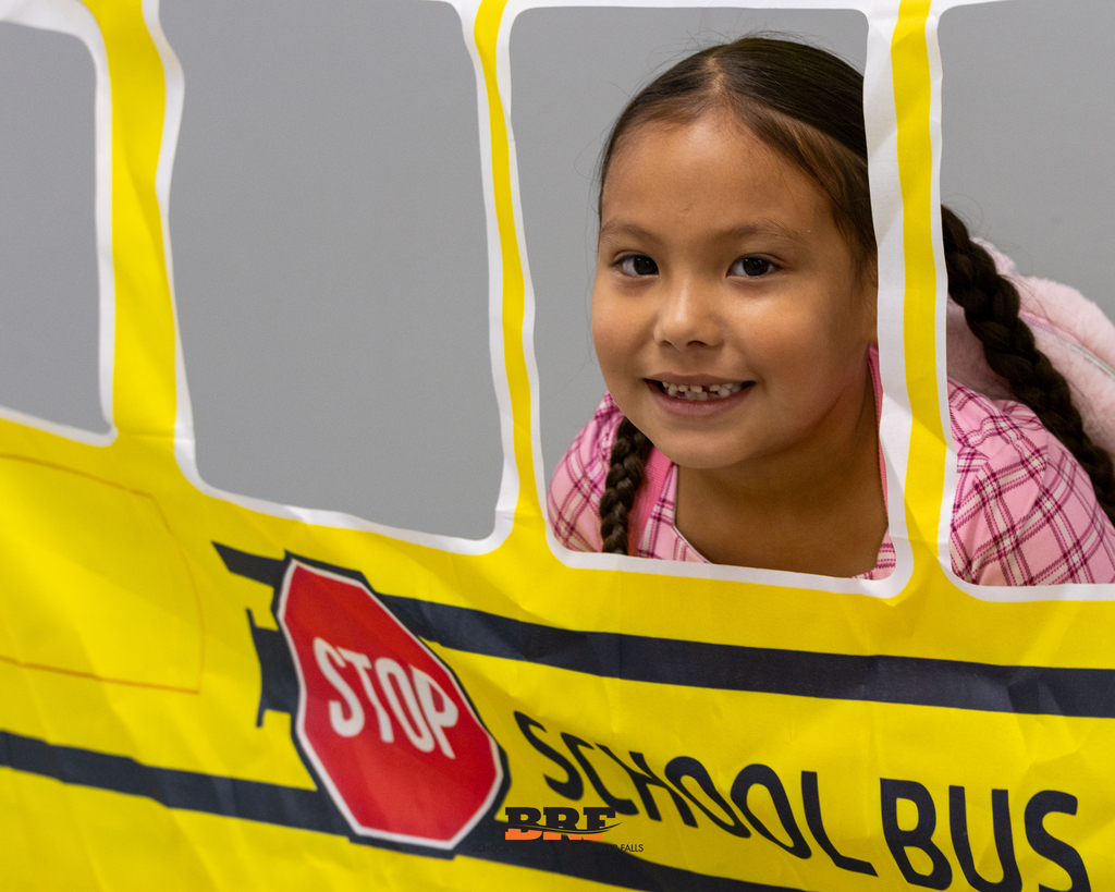 A young girl smiling happily while looking through the window cutout of a yellow school bus prop.