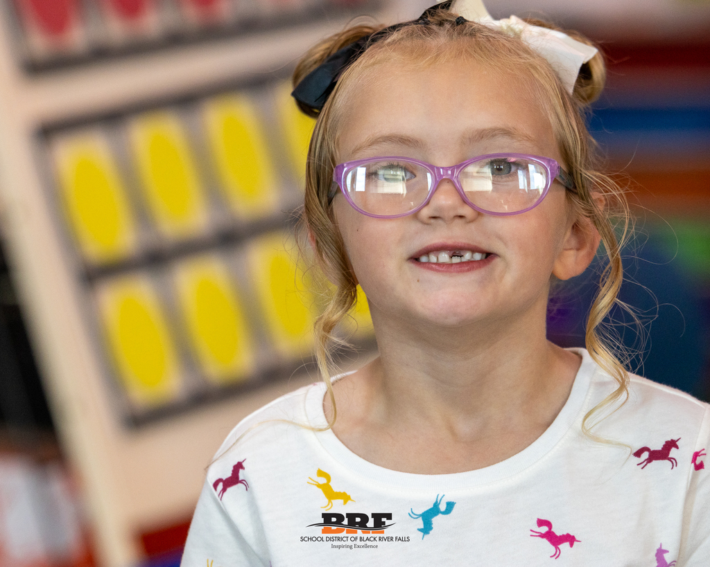 A young girl with blonde hair in pigtails, wearing purple glasses and a white unicorn shirt, smiling cheerfully for the camera.
