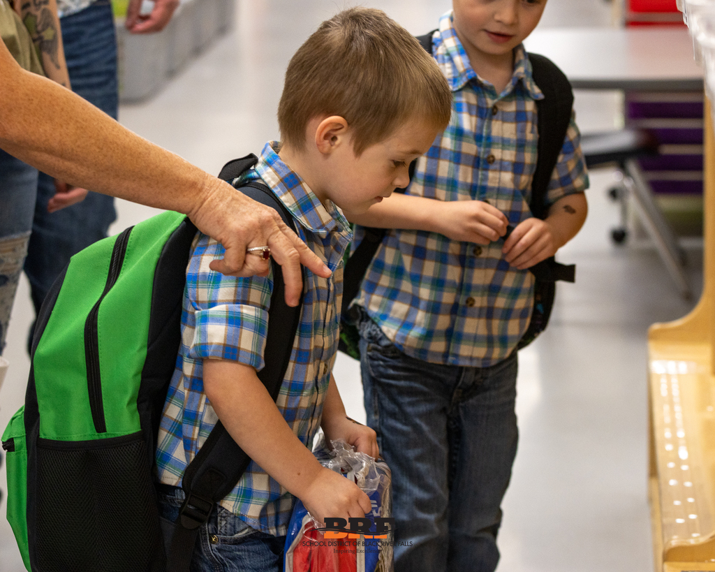 Two young students wearing matching blue plaid shirts and backpacks looking down while exploring classroom cubbies.