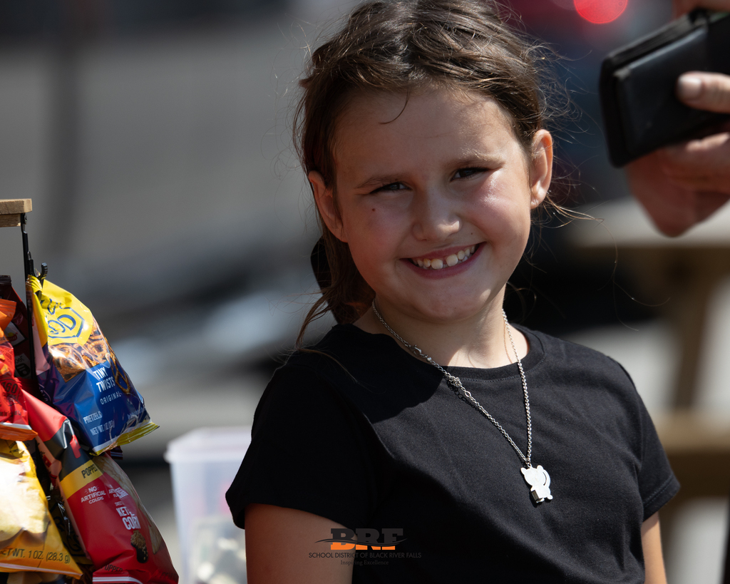 A young girl in a black t-shirt and silver necklace smiling brightly next to a rack of food truck snacks.