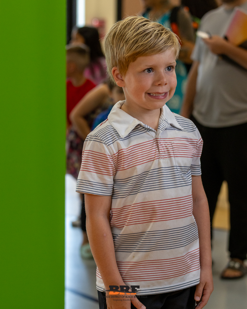 A young blonde boy in a striped polo shirt smiling cheerfully while standing near a bright green wall.