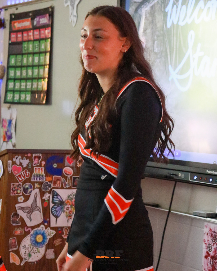 A high school cheerleader in a black and orange uniform smiles while standing near a whiteboard in a decorated classroom.