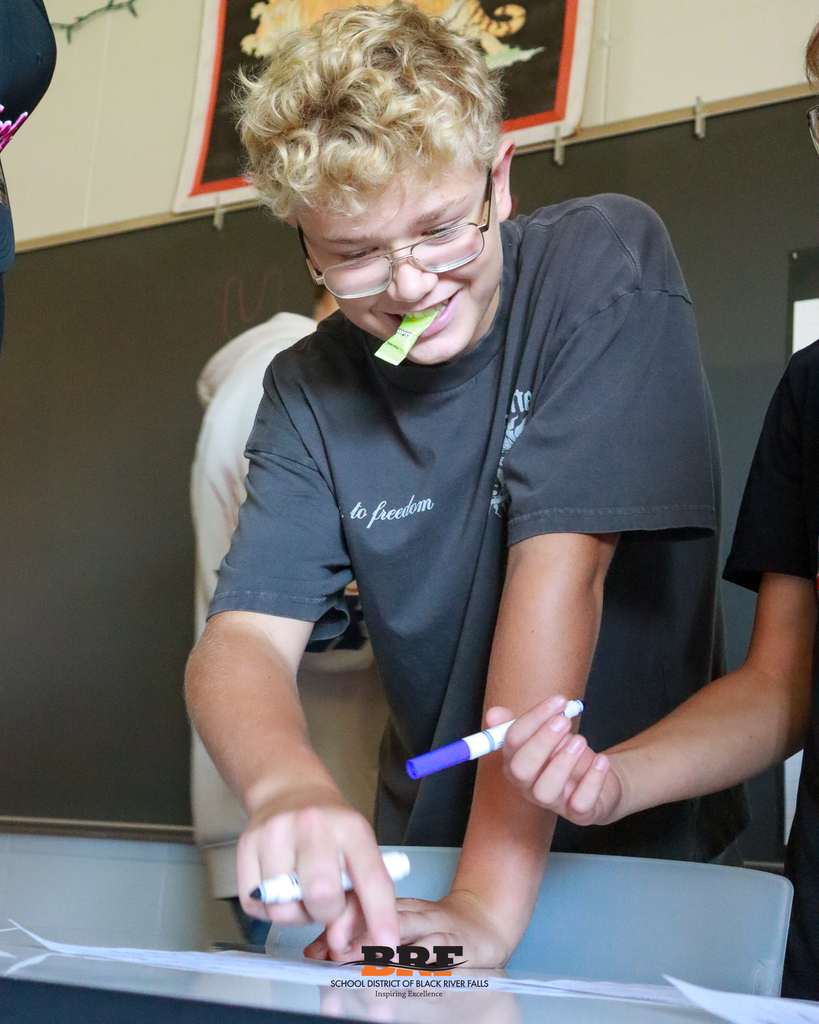 A blonde student wearing glasses smiles while holding dry-erase markers and writing on paper during a group activity.
