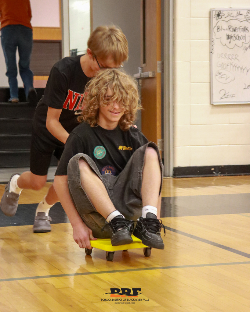 A boy with curly hair laughs as he sits on a yellow four-wheeled gym scooter while a classmate pushes him down a school hallway.