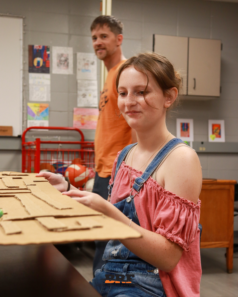 A high school student wearing a pink top and denim overalls smiles while working on a cardboard maze project in a classroom, with a teacher standing in the background.