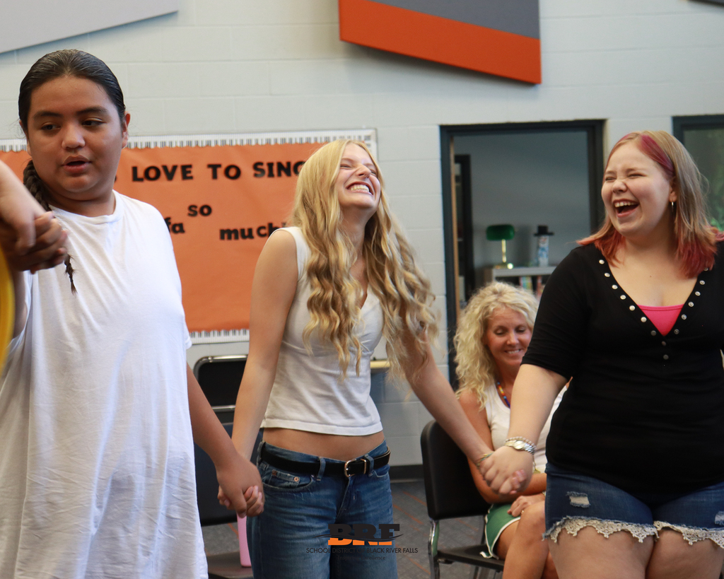 Three high school students hold hands in a circle and laugh together during a team-building activity in a classroom.