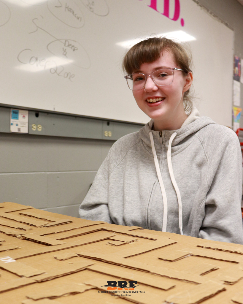A high school student with glasses and a grey hoodie smiles warmly at the camera while sitting in front of a cardboard maze.