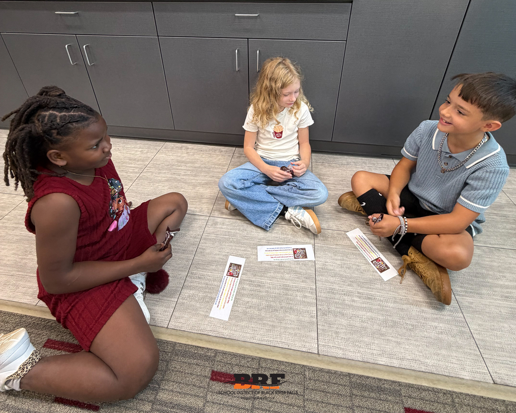 Three elementary students sit cross-legged on a classroom floor in a circle, holding small snacks and talking next to activity sheets.