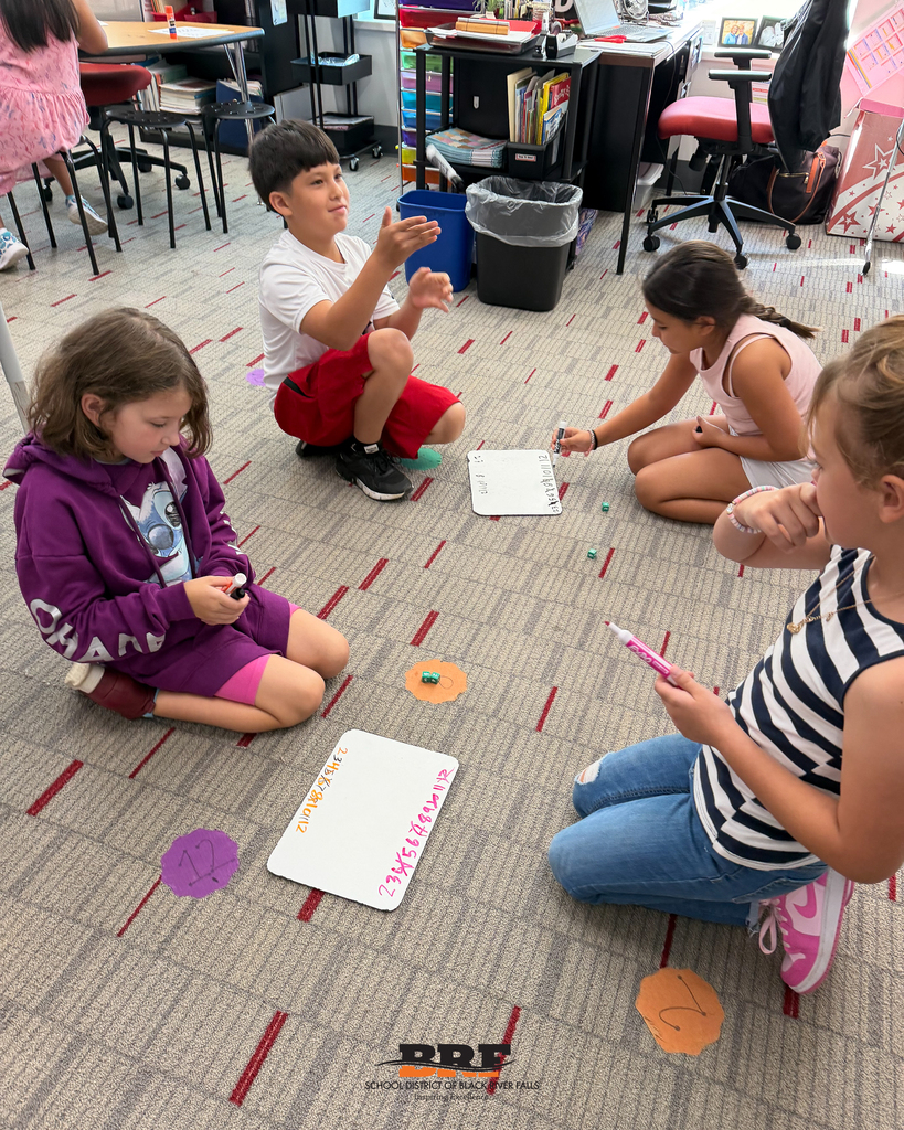 Four young students sit on the carpet around dry-erase boards, using markers to write out math problems during a group lesson.