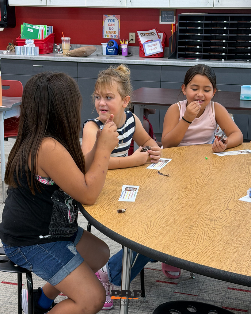 Three young students sit around a wooden classroom table, smiling and chatting while working on a hands-on learning activity.