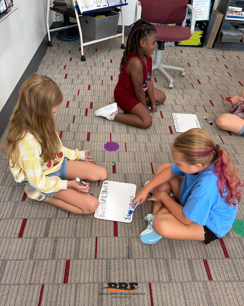 Three elementary students sit together on a classroom carpet, working together with dry-erase markers on a whiteboard activity.