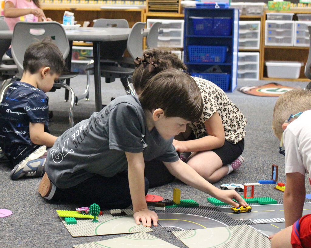 Two young elementary students sit on a classroom carpet, collaborating and driving toy cars along a colorful Lego road map during a hands-on activity.