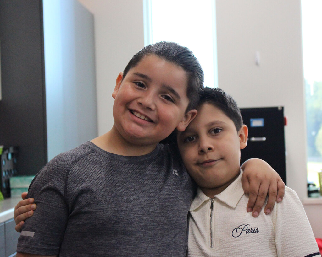 Two young boys stand close together in a classroom, smiling warmly at the camera with an arm wrapped around each other's shoulder.