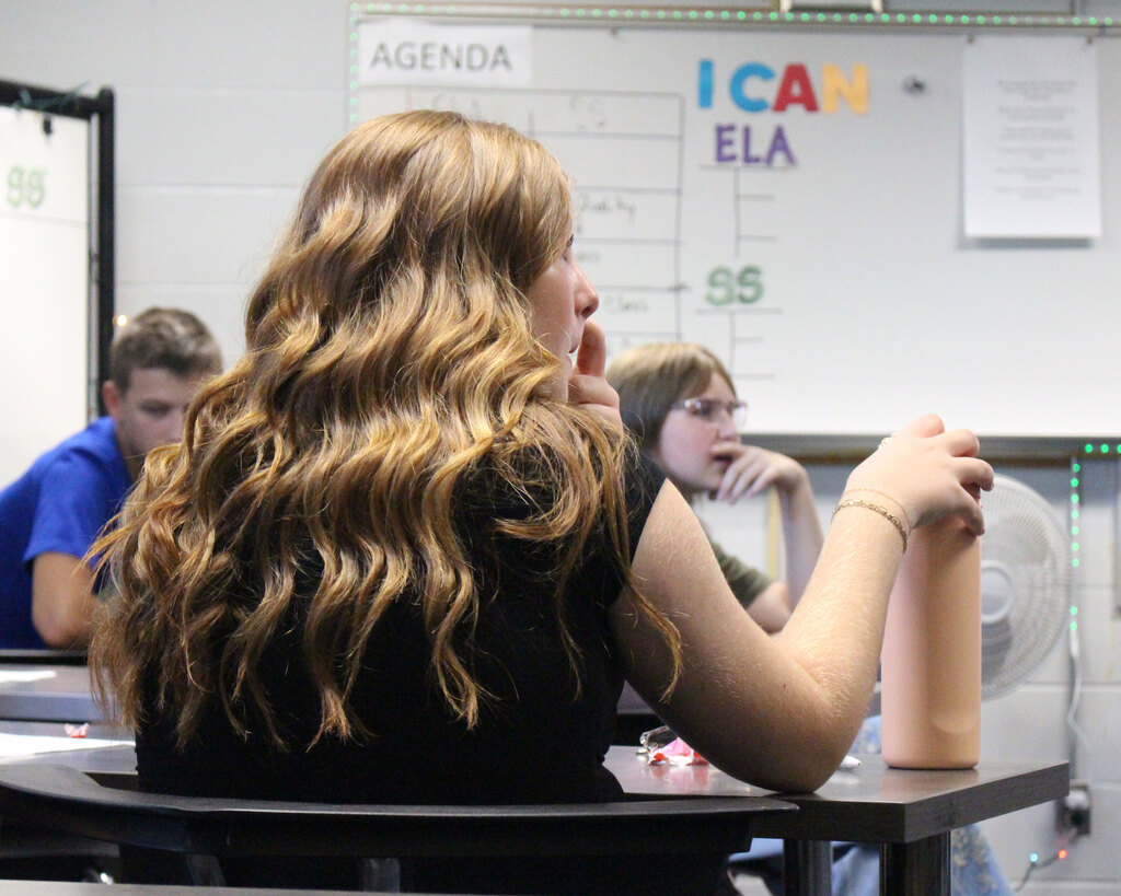 A middle school student with wavy blonde hair sits at her desk, attentively listening to a classroom discussion in front of a whiteboard reading "I CAN."