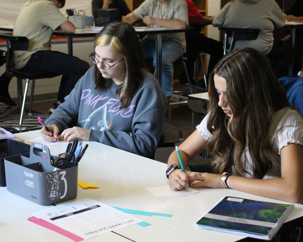 Two students sit side by side at a white classroom desk, focused on writing notes on index cards during a lesson.