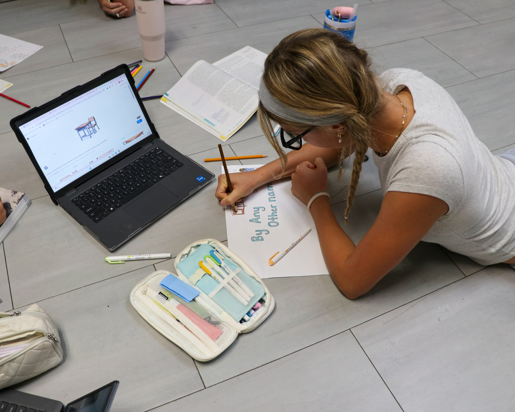 A student sitting on the floor leaning over a poster, using colored pencils to add fine details to a group assignment.