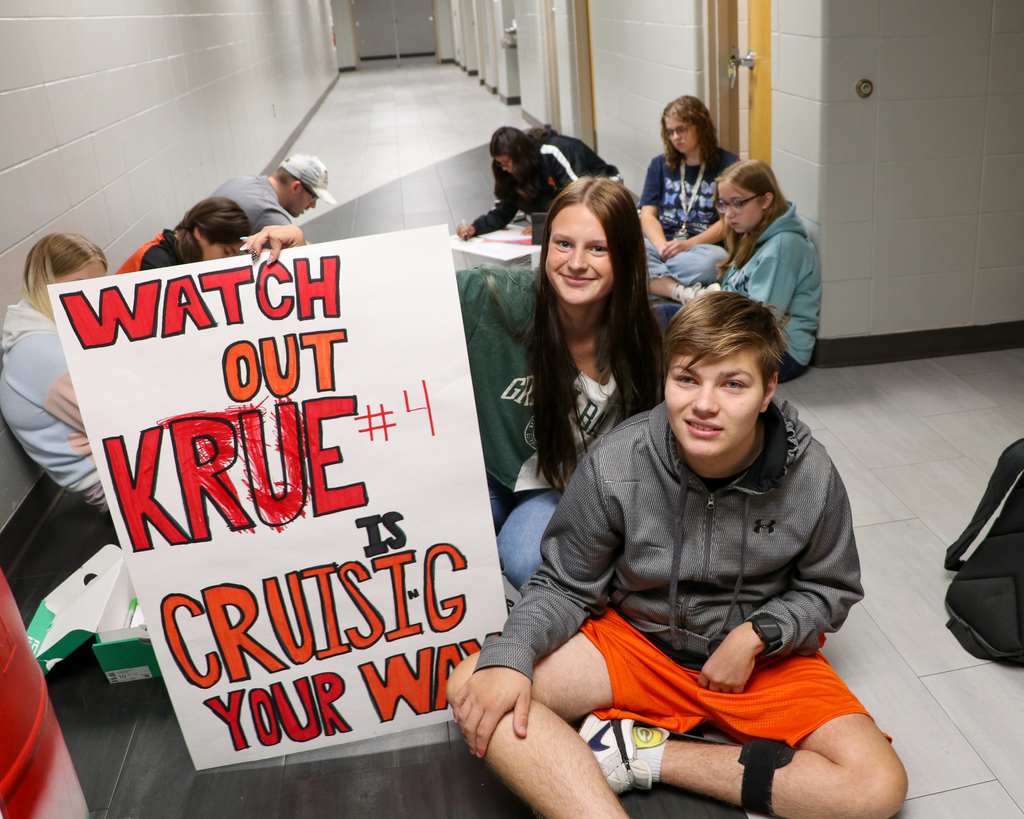 High school students kneeling on the floor around a poster, smiling as one student holds up a completed game-day spirit sign toward the camera.