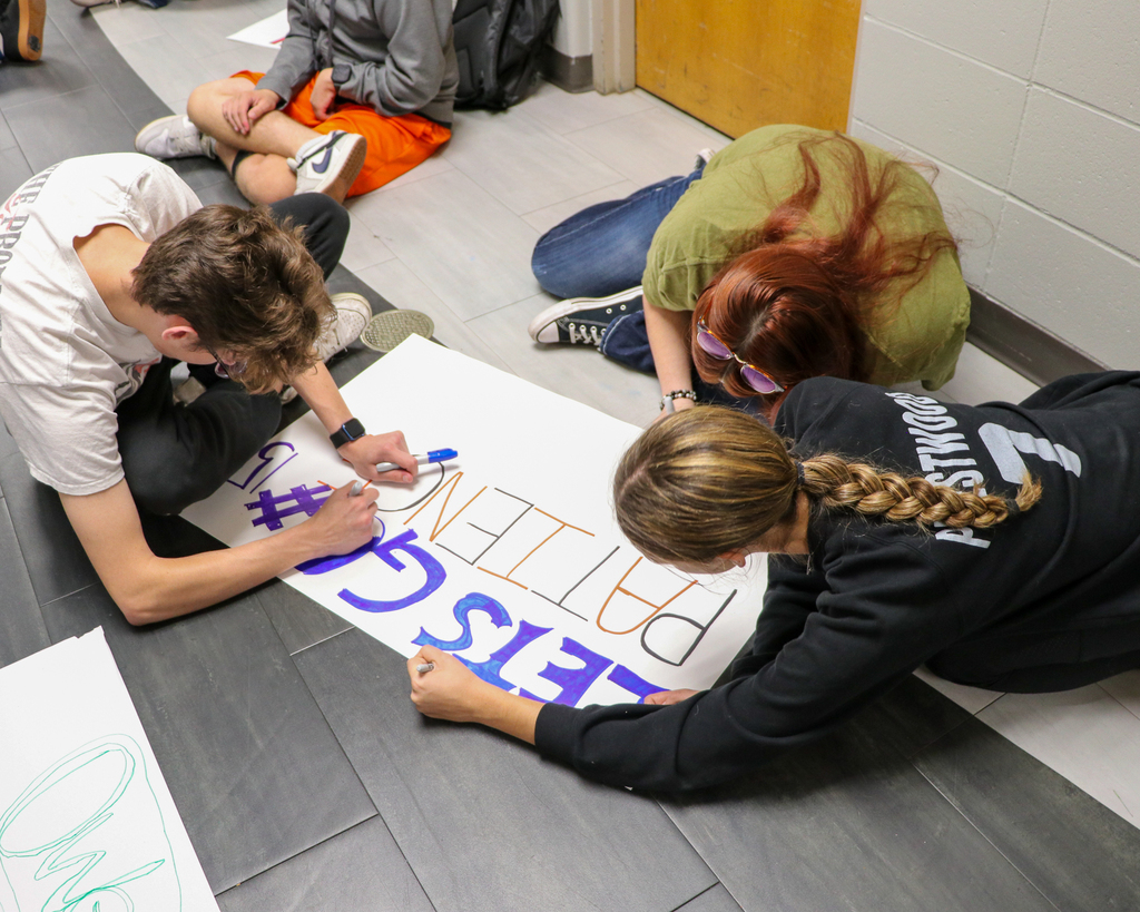 High school students sitting on the classroom floor around a large poster board, using markers to sketch out design concepts together.