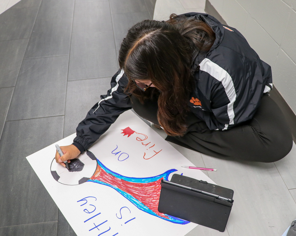 A high school student sitting cross-legged on the floor, focused on coloring and detailing a section of a poster project.