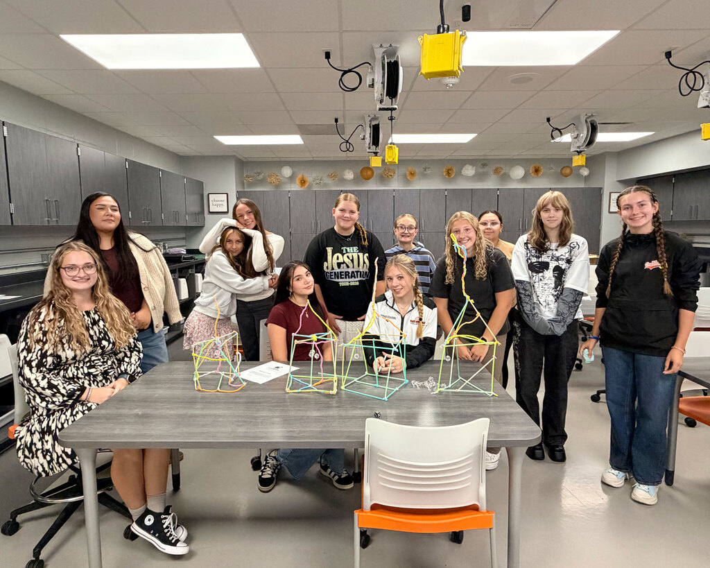 A group of high school students gather closely in a classroom around several detailed models they built. The students have used colorful pipe cleaners and plastic drinking straws to create intricate, network-like structures on tabletops and posters, representing neural connections in the human brain. The models vary in color, featuring blues, oranges, greens, and pinks.