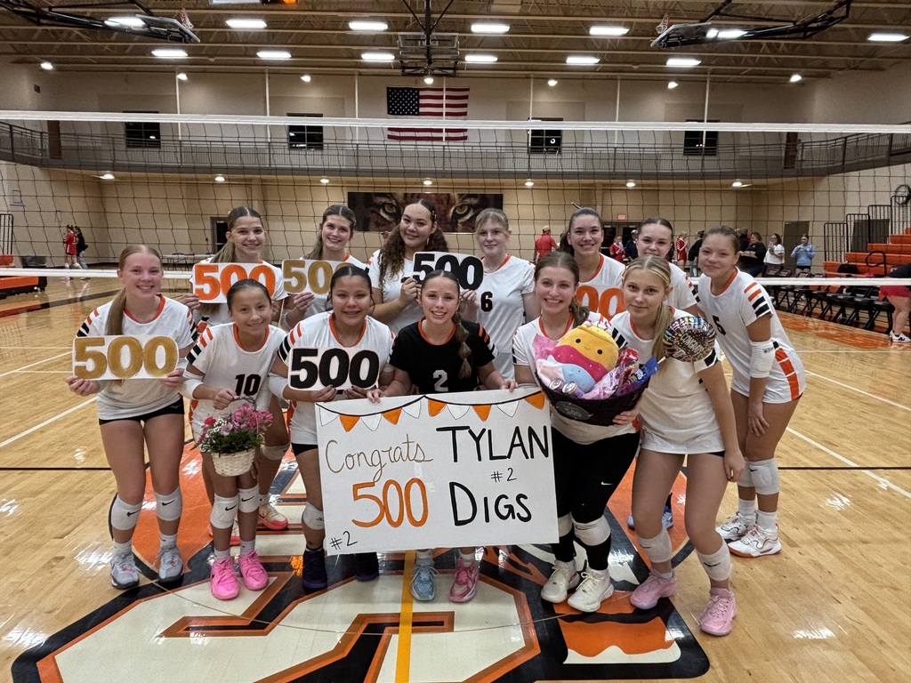 Tylan Brown poses on the volleyball court surrounded by her smiling teammates. The team holds up cutout numbers spelling out "500" along with a decorated sign that reads "Congrats Tylan #2 - 500 Digs" and a gift basket filled with treats.