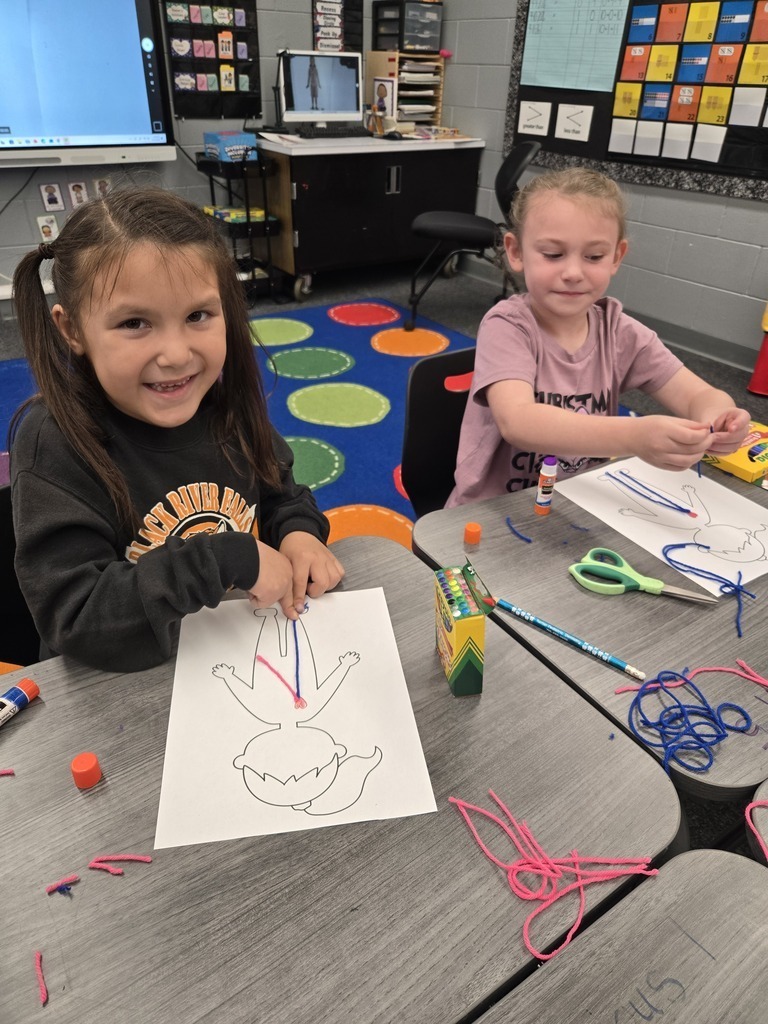 Two first-grade students sit side by side at classroom desks, smiling while gluing pieces of pink and blue yarn onto their human body outline worksheets.