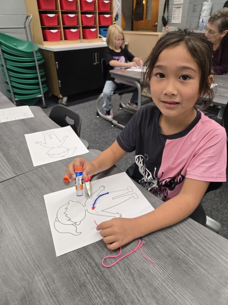 A first-grade student smiles at the camera while working at a grey classroom desk, carefully attaching pieces of blue and pink yarn to a human body outline using a glue stick.