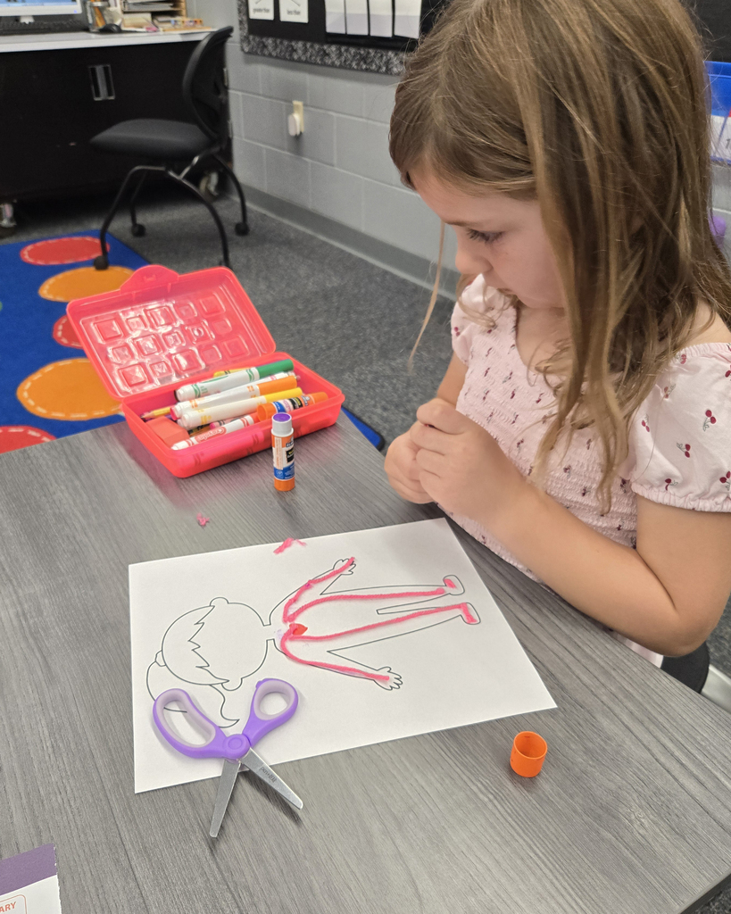 A first-grade student sits at a classroom table focused on gluing pink yarn onto a human body outline worksheet to represent the circulatory system. On the table sit a glue stick, a open pencil box with markers, and a pair of purple scissors.