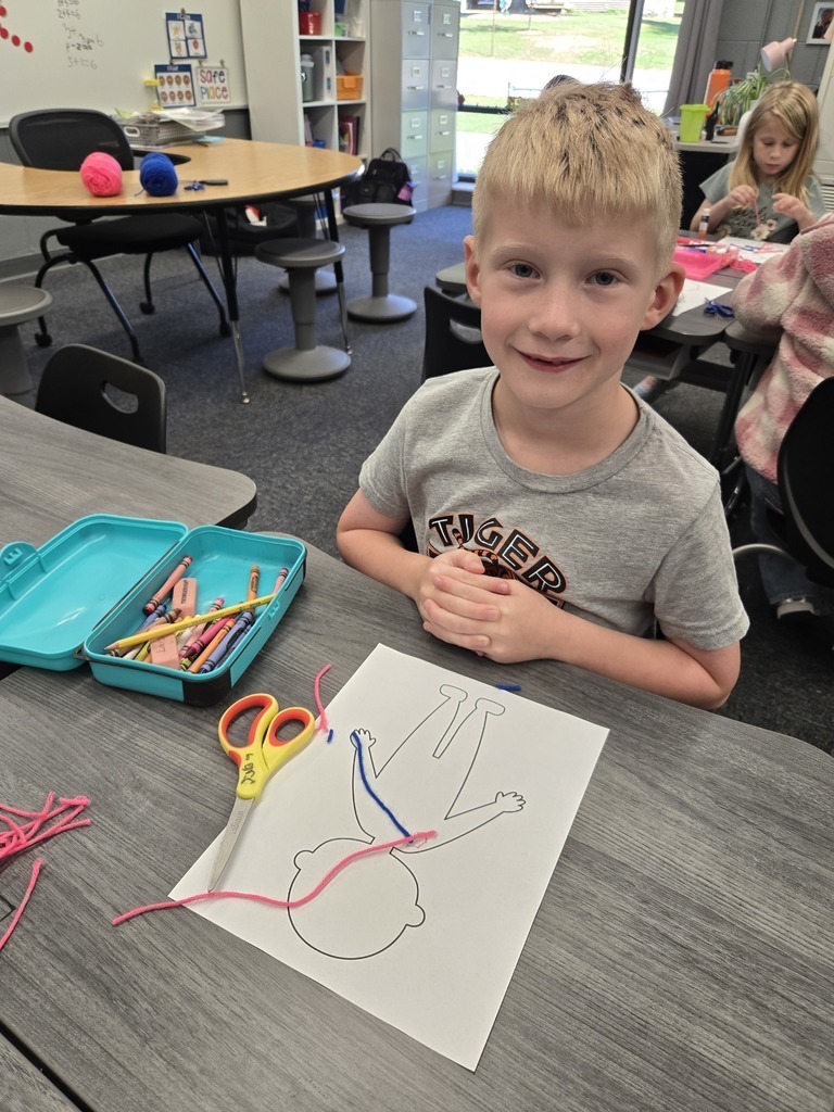 A first-grade student wearing a grey shirt that reads "Tiger" smiles for the camera at his desk, displaying his human body worksheet with blue and pink yarn representing blood vessels. A pencil box full of crayons and yellow scissors sit beside his paper.