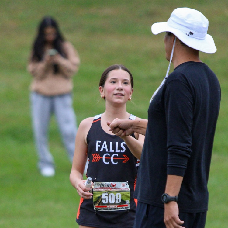 A high school female student-athlete, wearing a 'FALLS CC' black and orange singlet and bib number '509', speaks with an adult male coach, who wears a black long-sleeve shirt and a white bucket hat, at an outdoor cross-country meet. The coach stands with his back to the camera, gesturing with his hand. In the background, a person holds their phone in a grassy field.
