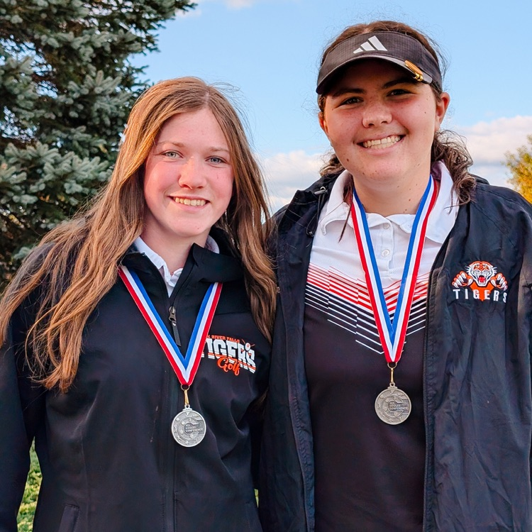 Two high school female student-athletes smile while wearing red, white, and blue medal ribbons around their necks. The student on the left wears a black jacket with 'Black River Falls Tigers' text and logo, and the student on the right wears a black jacket with 'Tigers' text and logo. Both students have medium-length brown hair.