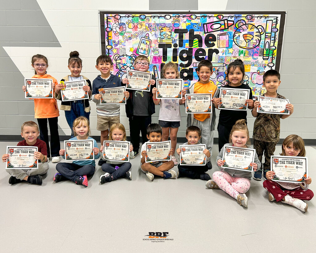 A group of young elementary students sitting and standing in two rows, smiling and proudly holding up "The Tiger Way" certificate awards in front of a colourful wall banner.