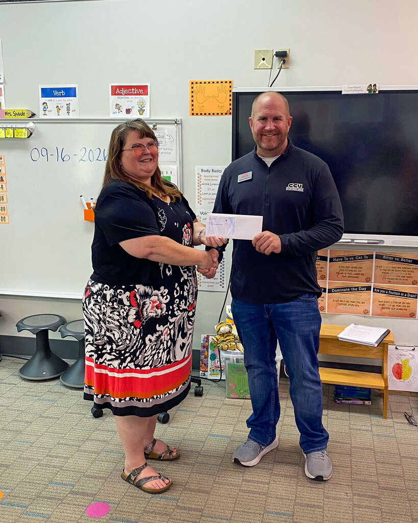 A woman in a patterned black and white dress and a man wearing a dark long-sleeved shirt and jeans smiling together in a classroom, joint-holding a white paper check. In the background are educational posters, a whiteboard, and classroom furniture.