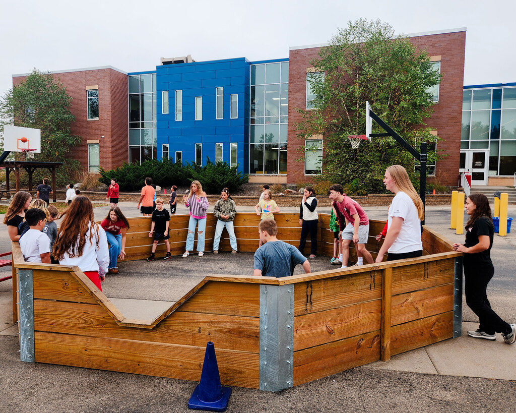 A group of students plays gaga ball inside an outdoor wooden octagonal pit on the school playground during an afternoon break. Students stand along the wooden perimeter watching and participating in the game in front of the school building.