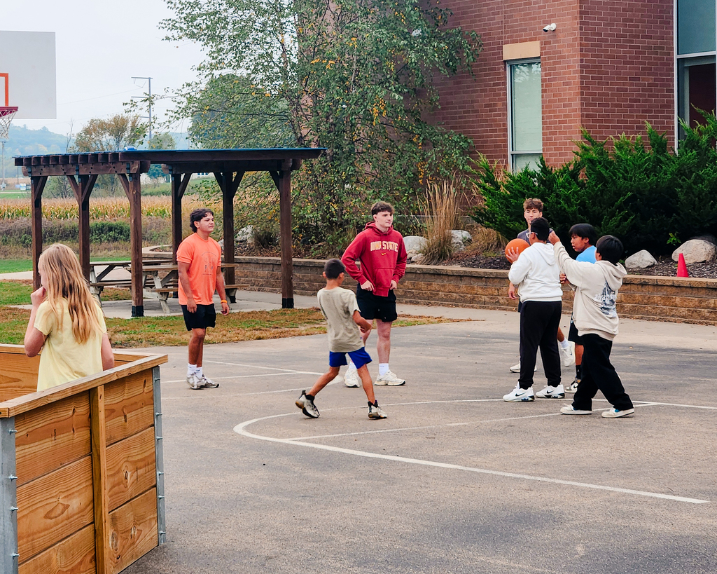 Students gather on an outdoor blacktop basketball court near a wooden gaga pit during bonus recess. A group of students plays basketball while others look on near the playground area.