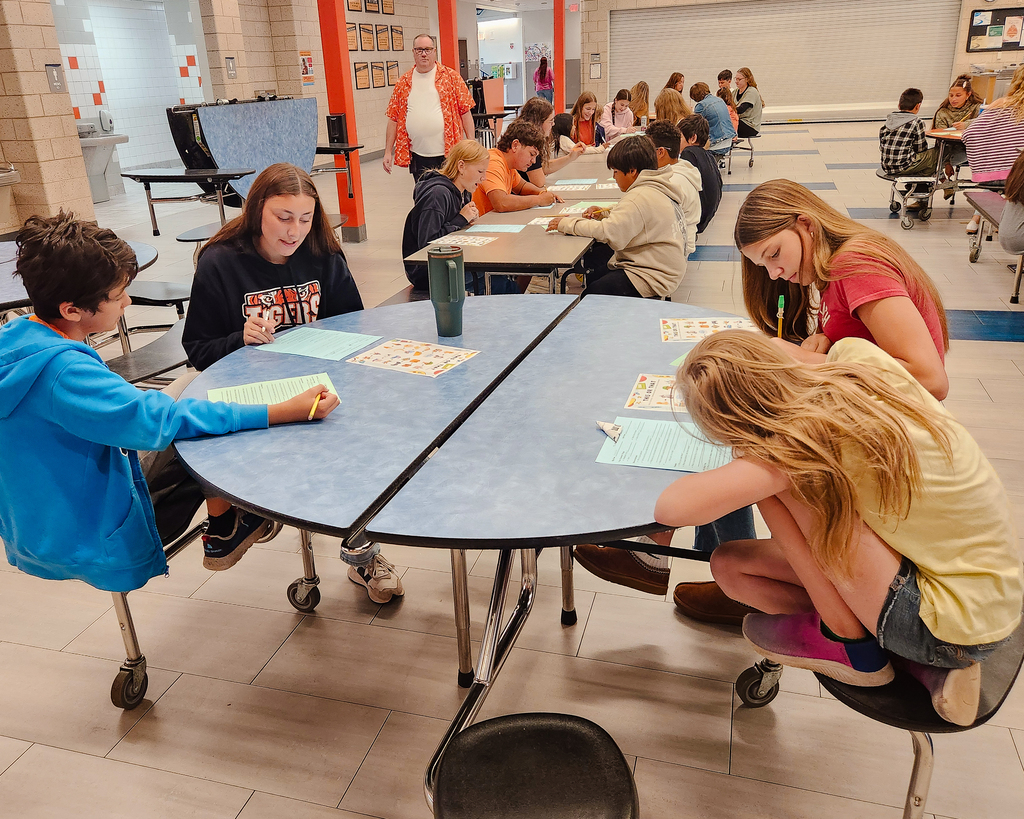 Students sit paired together at round blue tables in the school commons working on printed Big Paw/Little Paw interview and matching worksheets. One student leans over the table writing answers while working alongside a mentor partner.