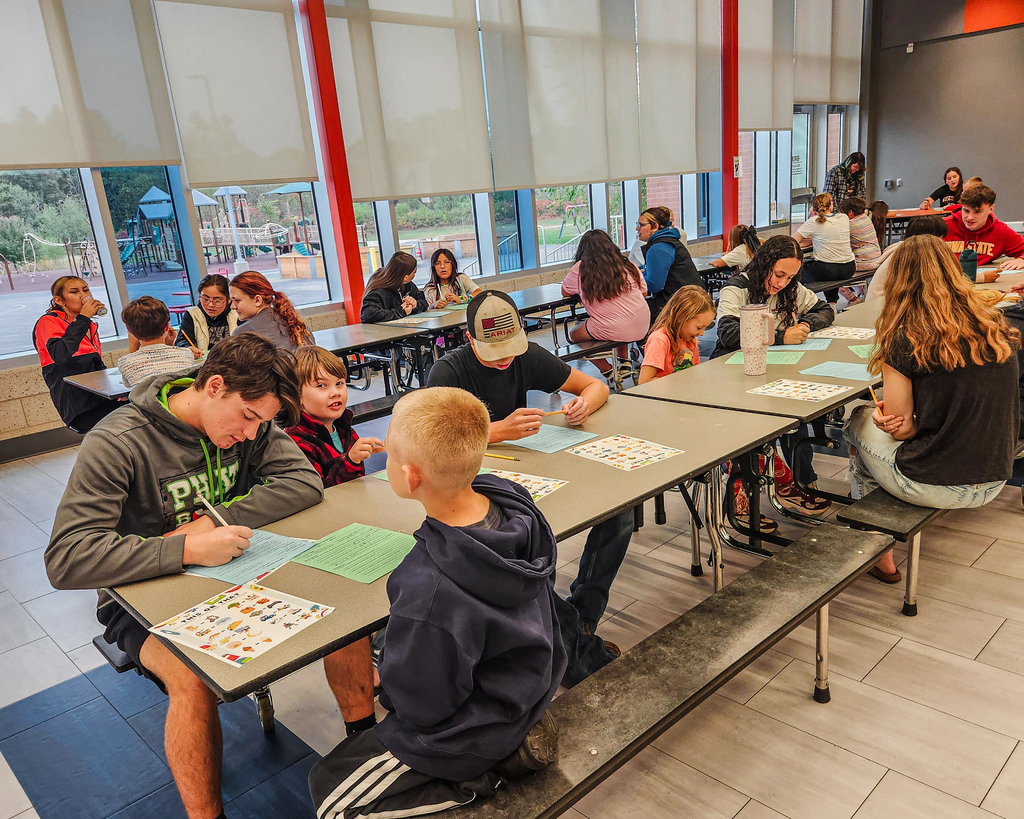 Students sit together along long cafeteria tables during a Big Paw/Little Paw pairing activity. Students write on green "This or That" worksheets and talk with their designated partners.