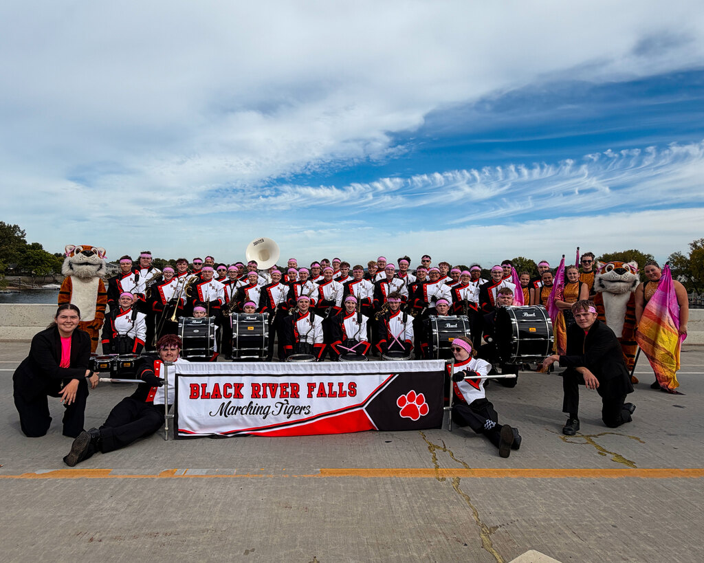 Members of the Black River Falls Marching Tigers posing together outdoors with their banner and instruments.